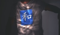 A person is wearing a light-colored shirt with a large blue square on the front, containing a white Asian character. Below the square, the word 'PEACE' is printed in capital letters. The shirt is partially in shadow, creating an interesting pattern of light and dark.