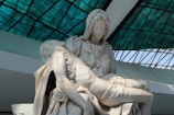 A marble sculpture depicting two figures, with one figure cradling another in a tender and sorrowful manner. The backdrop features a grid-like pattern created by a structure with green stained glass panels.