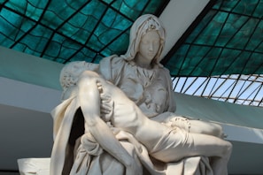A marble sculpture depicting two figures, with one figure cradling another in a tender and sorrowful manner. The backdrop features a grid-like pattern created by a structure with green stained glass panels.