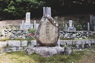 A large stone monument with engraved text stands in a serene, outdoor setting. It is surrounded by moss-covered ground, small plants, and other commemorative stones. The background features a stone wall and a densely wooded area, creating a tranquil and solemn atmosphere.