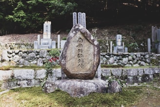A large stone monument with engraved text stands in a serene, outdoor setting. It is surrounded by moss-covered ground, small plants, and other commemorative stones. The background features a stone wall and a densely wooded area, creating a tranquil and solemn atmosphere.