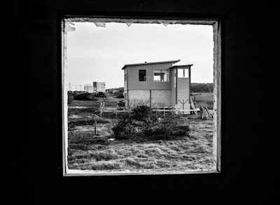 A house sits in the center of a grassy landscape, framed by a large, dark window in the foreground. The structure appears to be a modern, two-story building with horizontal siding and a slight overhang on the roof. There's a field with grass and small bushes surrounding the house, providing a stark, open contrast to the enclosed frame.