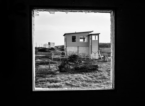 A house sits in the center of a grassy landscape, framed by a large, dark window in the foreground. The structure appears to be a modern, two-story building with horizontal siding and a slight overhang on the roof. There's a field with grass and small bushes surrounding the house, providing a stark, open contrast to the enclosed frame.