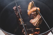 Several pieces of meat, including sausages and ribs, are hanging inside a metal smoker above a fire, showing a rustic cooking process.