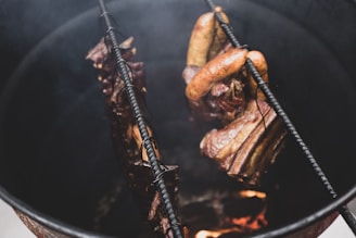 Several pieces of meat, including sausages and ribs, are hanging inside a metal smoker above a fire, showing a rustic cooking process.