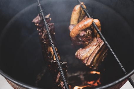 Several pieces of meat, including sausages and ribs, are hanging inside a metal smoker above a fire, showing a rustic cooking process.