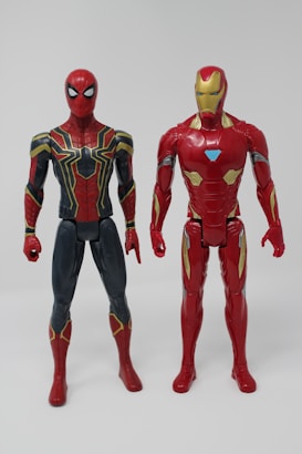 Two action figures of superheroes stand side by side against a plain background. The figure on the left is dressed in a red and navy suit with intricate gold detailing, while the figure on the right is clad in a predominantly red metallic suit with gold and blue accents.