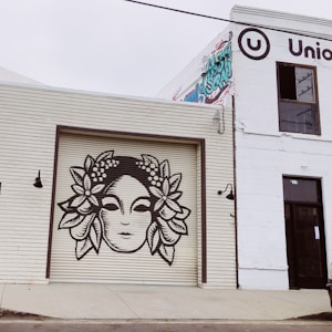 A mural of a stylized face with flowers in the hair painted on the roll-up door of a building. The building next to it has the word 'Union' partially visible with graffiti art on the wall.