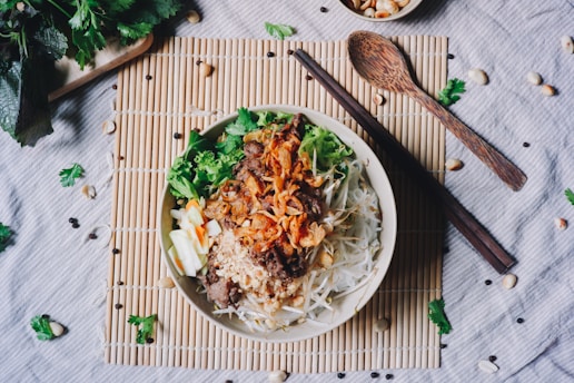 A bowl filled with a colorful and appetizing meal, including vegetables, meat, crispy fried onions, and possibly noodles or sprouts. Surrounding the bowl are various garnishes, fresh herbs, and a wooden spoon and chopsticks placed on a bamboo mat. The setting is casual and inviting, with a rustic feel enhanced by the scattered ingredients and the wooden utensils.