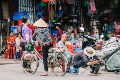 A bustling street scene features a group of people engaged in various activities. One person is working on a scooter, while another person is standing with a bicycle. A vendor's stall in the background displays an array of goods, including colorful plastic chairs and other household items. People are casually gathered, wearing everyday attire, and one of them is wearing a traditional conical hat.