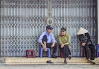 Three elderly individuals are sitting on small plastic stools in front of a large metal gate. Two men, wearing caps, are engaged in conversation while a woman, wearing a traditional conical hat, sits with her head down. The scene conveys a sense of quiet companionship and everyday life.