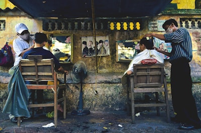 An outdoor barbershop setup along a street with two barbers cutting the hair of two customers seated on wooden chairs. The background consists of a weathered stone wall with mirrors and posters affixed to it. One barber is wearing a cap and a face mask, and the other is similarly masked. A standing fan and scattered tools are visible on the ground.