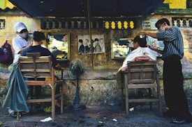 An outdoor barbershop setup along a street with two barbers cutting the hair of two customers seated on wooden chairs. The background consists of a weathered stone wall with mirrors and posters affixed to it. One barber is wearing a cap and a face mask, and the other is similarly masked. A standing fan and scattered tools are visible on the ground.