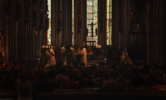 A church interior with stained glass windows in the background, where a religious ceremony is taking place. Several people dressed in liturgical robes stand near an altar decorated with candles and religious symbols, while a congregation is seated observing the ceremony.