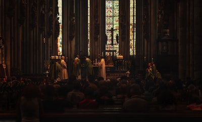 A church interior with stained glass windows in the background, where a religious ceremony is taking place. Several people dressed in liturgical robes stand near an altar decorated with candles and religious symbols, while a congregation is seated observing the ceremony.