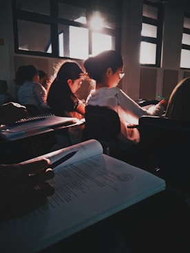 Students sit in a dimly lit classroom, focused on their work with sunlight streaming through the windows creating a warm glow. Papers and open books are visible, suggesting a study or lecture setting.