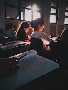 Students sit in a dimly lit classroom, focused on their work with sunlight streaming through the windows creating a warm glow. Papers and open books are visible, suggesting a study or lecture setting.
