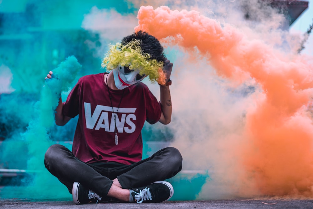 face painted man sitting on ground surrounded by orange and green smoke