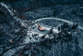 Heavy construction equipment sits on a winding dirt road within a rocky quarry. The machinery is surrounded by rugged rock walls, and the foreground features blurry autumn leaves, adding depth to the scene.