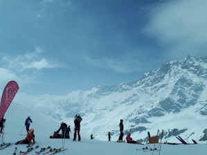 A snowy mountain landscape with several people engaged in skiing activities. The scene includes multiple skiers equipped with gear such as ski poles and helmets. A tall mountain range with snow-covered peaks provides a dramatic backdrop, while the sky is mostly cloudy with patches of blue. A branded flag is visible on the left, possibly indicating a ski resort or location.