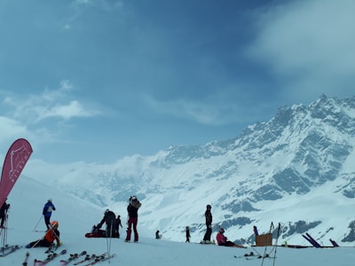 A snowy mountain landscape with several people engaged in skiing activities. The scene includes multiple skiers equipped with gear such as ski poles and helmets. A tall mountain range with snow-covered peaks provides a dramatic backdrop, while the sky is mostly cloudy with patches of blue. A branded flag is visible on the left, possibly indicating a ski resort or location.