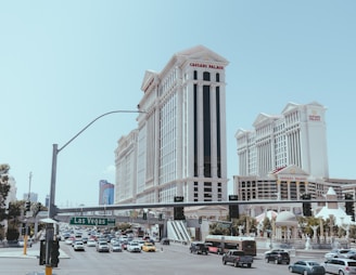 A bustling street scene with heavy traffic and prominent buildings. The architecture features a grand style, with tall, ornate structures like Caesars Palace. Vehicles of various types are seen on the road, and a green street sign indicates Las Vegas Boulevard. There is a clear sky in the background.