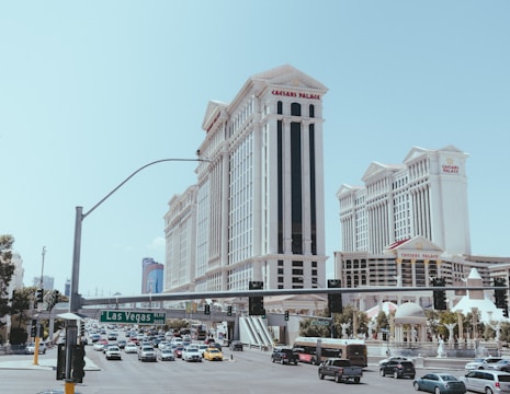 A bustling street scene with heavy traffic and prominent buildings. The architecture features a grand style, with tall, ornate structures like Caesars Palace. Vehicles of various types are seen on the road, and a green street sign indicates Las Vegas Boulevard. There is a clear sky in the background.
