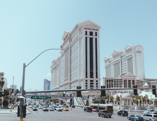 A bustling street scene with heavy traffic and prominent buildings. The architecture features a grand style, with tall, ornate structures like Caesars Palace. Vehicles of various types are seen on the road, and a green street sign indicates Las Vegas Boulevard. There is a clear sky in the background.