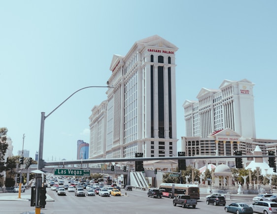 A bustling street scene with heavy traffic and prominent buildings. The architecture features a grand style, with tall, ornate structures like Caesars Palace. Vehicles of various types are seen on the road, and a green street sign indicates Las Vegas Boulevard. There is a clear sky in the background.