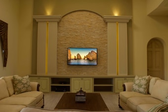 A cozy living room features a wall-mounted television displaying a scenic image of rock formations by the sea. The TV is framed by a decorative stone wall and flanked by two classical columns with recessed lighting. Two beige sofas with patterned cushions are positioned around a dark wooden coffee table. The room has a soothing, warm color palette and elegant furnishings.