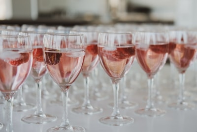 Rows of elegant wine glasses containing a pink liquid with ice cubes and pieces of fruit are neatly arranged on a white surface.