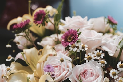A bouquet featuring a mix of flowers including pale pink roses, purple asters, white daisies, and creamy yellow lilies, arranged with green foliage.