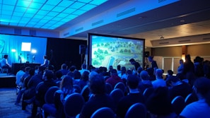 A group of people are seated in a dimly lit room, facing a large screen displaying a colorful image of a landscape, possibly from a video game. The room has a blue lighting theme and a modern ceiling design. Some individuals are standing and observing near the back and sides of the room. On the left side, there are two people with a setup that includes monitors and a camera.