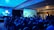 A group of people are seated in a dimly lit room, facing a large screen displaying a colorful image of a landscape, possibly from a video game. The room has a blue lighting theme and a modern ceiling design. Some individuals are standing and observing near the back and sides of the room. On the left side, there are two people with a setup that includes monitors and a camera.