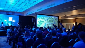 A group of people are seated in a dimly lit room, facing a large screen displaying a colorful image of a landscape, possibly from a video game. The room has a blue lighting theme and a modern ceiling design. Some individuals are standing and observing near the back and sides of the room. On the left side, there are two people with a setup that includes monitors and a camera.