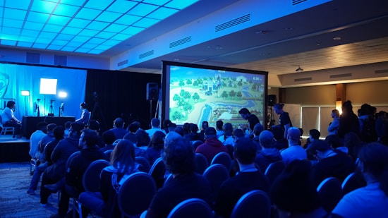 A group of people are seated in a dimly lit room, facing a large screen displaying a colorful image of a landscape, possibly from a video game. The room has a blue lighting theme and a modern ceiling design. Some individuals are standing and observing near the back and sides of the room. On the left side, there are two people with a setup that includes monitors and a camera.