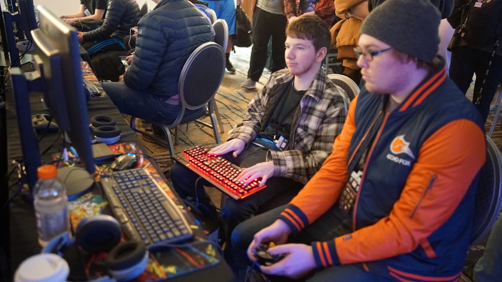 컴퓨터 키보드를 사용하는 남자 옆에 비디오 게임 컨트롤 패드를 사용하는 남자가 앉아서 비디오 게임을 하고 있습니다.