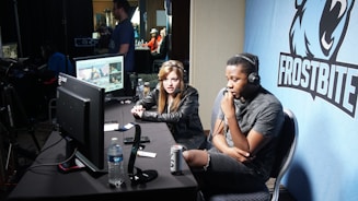 Two people are seated at a table with gaming monitors, wearing headsets. They appear to be participating in an esports broadcast or commentary. The environment is dimly lit with additional equipment and people visible in the background. A large blue banner with the word 'Frostbite' and a stylized graphic of a wolf is displayed.