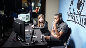Two people are seated at a table with gaming monitors, wearing headsets. They appear to be participating in an esports broadcast or commentary. The environment is dimly lit with additional equipment and people visible in the background. A large blue banner with the word 'Frostbite' and a stylized graphic of a wolf is displayed.