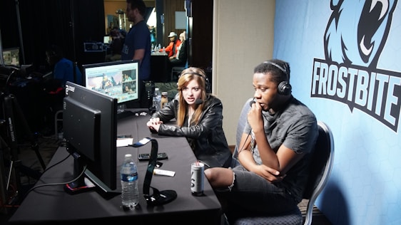 Two people are seated at a table with gaming monitors, wearing headsets. They appear to be participating in an esports broadcast or commentary. The environment is dimly lit with additional equipment and people visible in the background. A large blue banner with the word 'Frostbite' and a stylized graphic of a wolf is displayed.