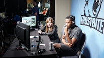 Two people are seated at a table with gaming monitors, wearing headsets. They appear to be participating in an esports broadcast or commentary. The environment is dimly lit with additional equipment and people visible in the background. A large blue banner with the word 'Frostbite' and a stylized graphic of a wolf is displayed.