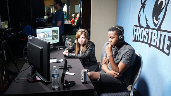 Two people are seated at a table with gaming monitors, wearing headsets. They appear to be participating in an esports broadcast or commentary. The environment is dimly lit with additional equipment and people visible in the background. A large blue banner with the word 'Frostbite' and a stylized graphic of a wolf is displayed.