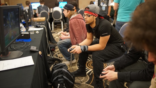 Several people are sitting in a room with gaming consoles and monitors, focusing intently on their screens. The setting appears to be a casual gaming event, with participants holding controllers while playing video games. The room has a casual atmosphere with chairs and tables set up for gaming.