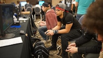 Several people are sitting in a room with gaming consoles and monitors, focusing intently on their screens. The setting appears to be a casual gaming event, with participants holding controllers while playing video games. The room has a casual atmosphere with chairs and tables set up for gaming.
