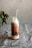 A glass bottle filled with a layered chocolate and cream beverage, topped with a straw, sits on a textured gray surface. A blurred green plant is visible in the background.