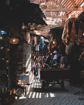 A bustling market scene with people walking through narrow pathways. Colorful fabrics and clothing are displayed alongside traditional crafts and ornaments. Light filters through wooden slats above, creating patterns on the cobblestone floor. A cart is being pushed by a person in the foreground.