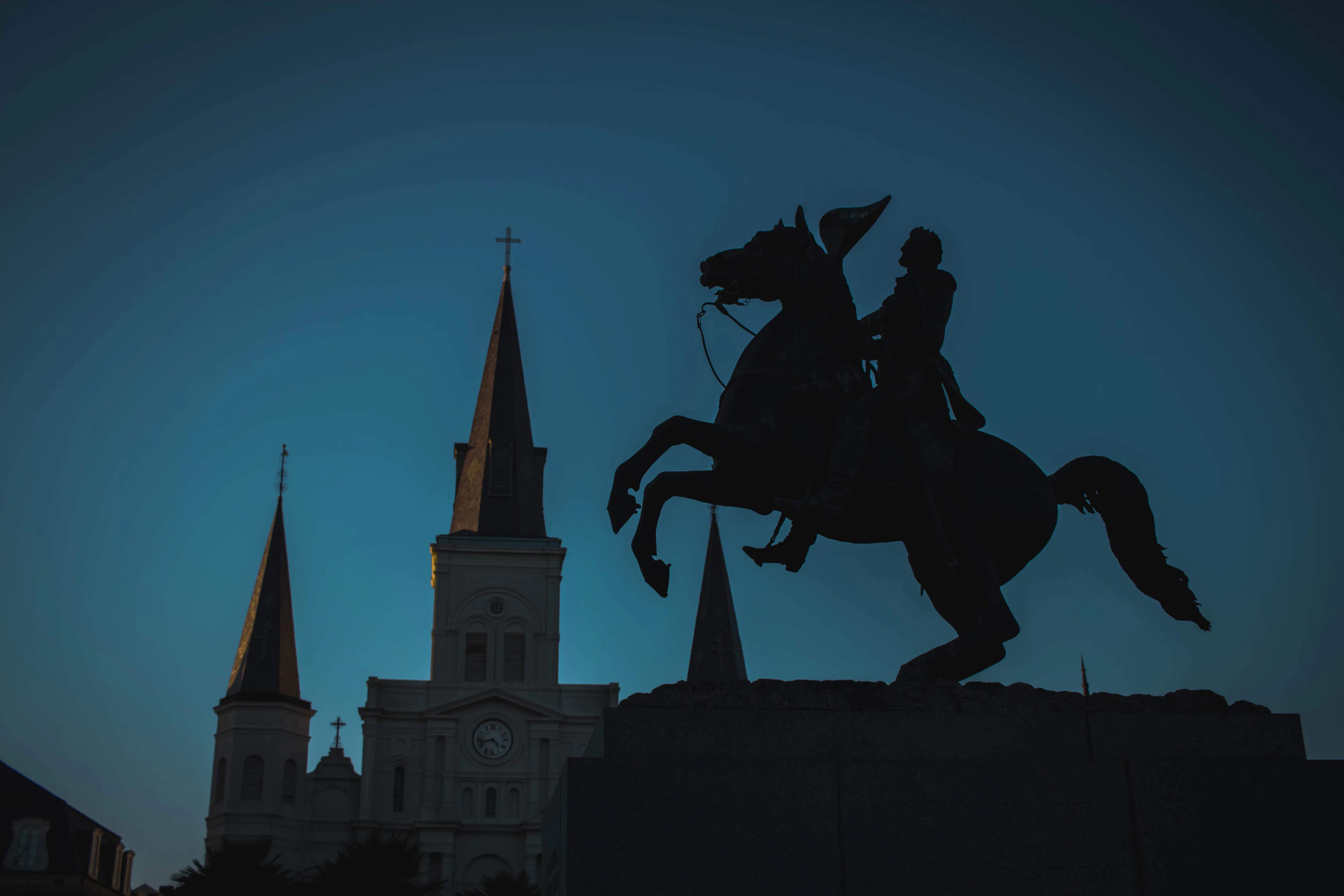 The bronze equestrian statue of Andrew Jackson silhouetted against a deep blue dusk sky in Jackson Square, New Orleans, with the three lit spires of St. Louis Cathedral rising behind it.