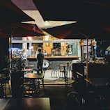 A cozy café or restaurant setting is captured at night. Several tables and chairs are present outside under large umbrellas, with some greenery in planters. The interior is warmly lit, and the view includes a bar area. Two people are standing near the counter, possibly placing orders or interacting with staff. The atmosphere is inviting and relaxed.