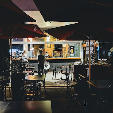 A cozy café or restaurant setting is captured at night. Several tables and chairs are present outside under large umbrellas, with some greenery in planters. The interior is warmly lit, and the view includes a bar area. Two people are standing near the counter, possibly placing orders or interacting with staff. The atmosphere is inviting and relaxed.