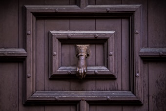 An ornate metallic door knocker shaped like a hand is mounted on a dark wooden door, which features a decorative square panel with rivets.